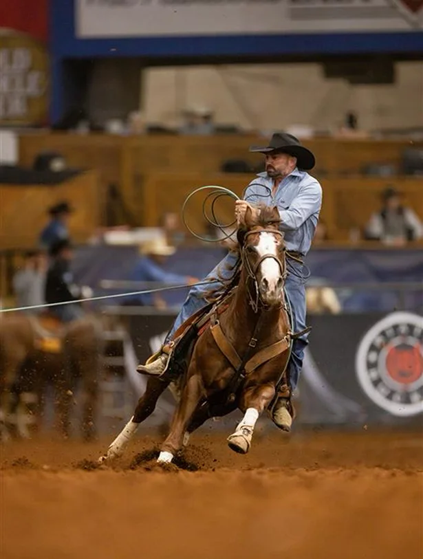 A cowboy in a hat rides Pepinstep in an indoor arena with a lasso