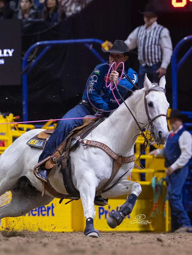 A cowboy in a black hat rides a SEVS Judgement Day at speed in an indoor rodeo arena, swinging a pink lasso