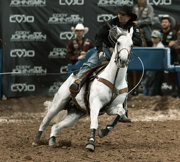 A cowboy in a black hat rides a SEVS Judgement Day at speed in an indoor rodeo arena, swinging a lasso
